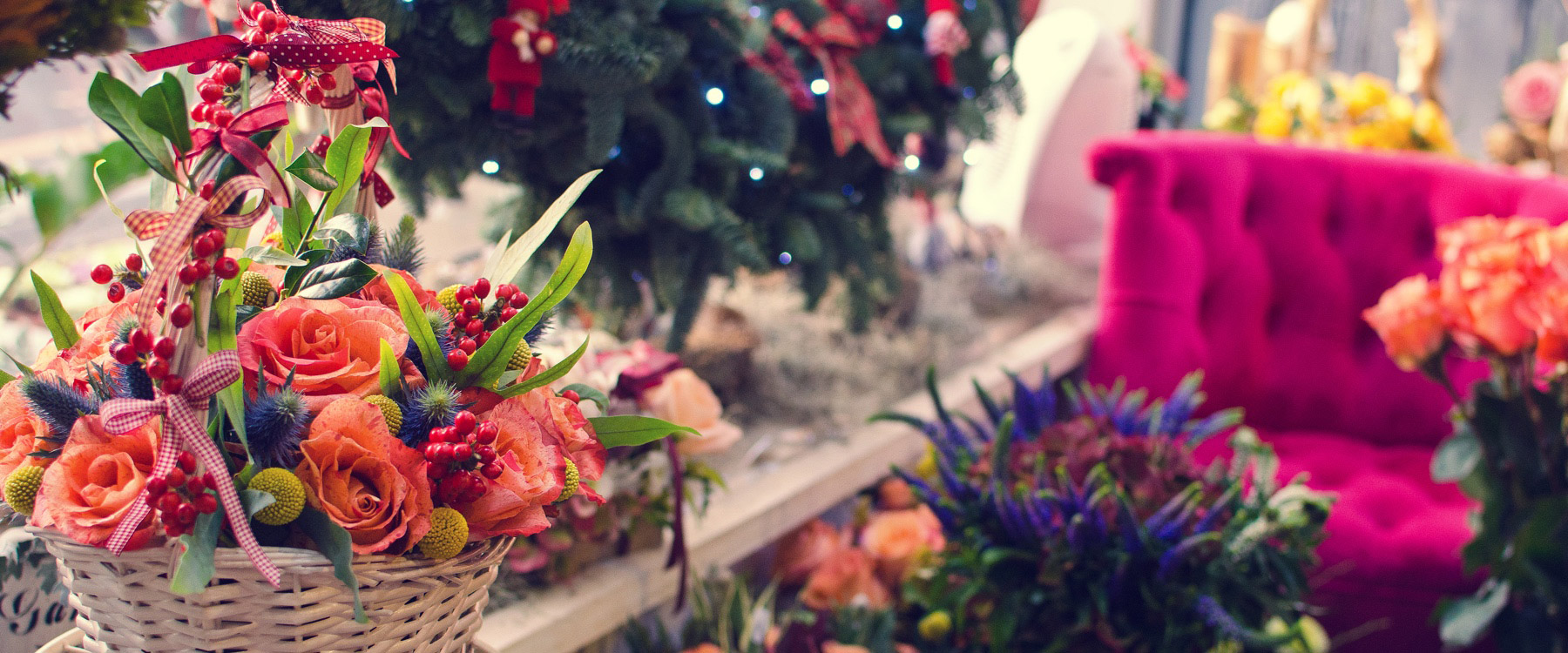 A white wicker basket filled with vibrant orange roses and red berries is set on a table near a decorated Christmas tree and a plush, pink chair in a festive indoor setting.