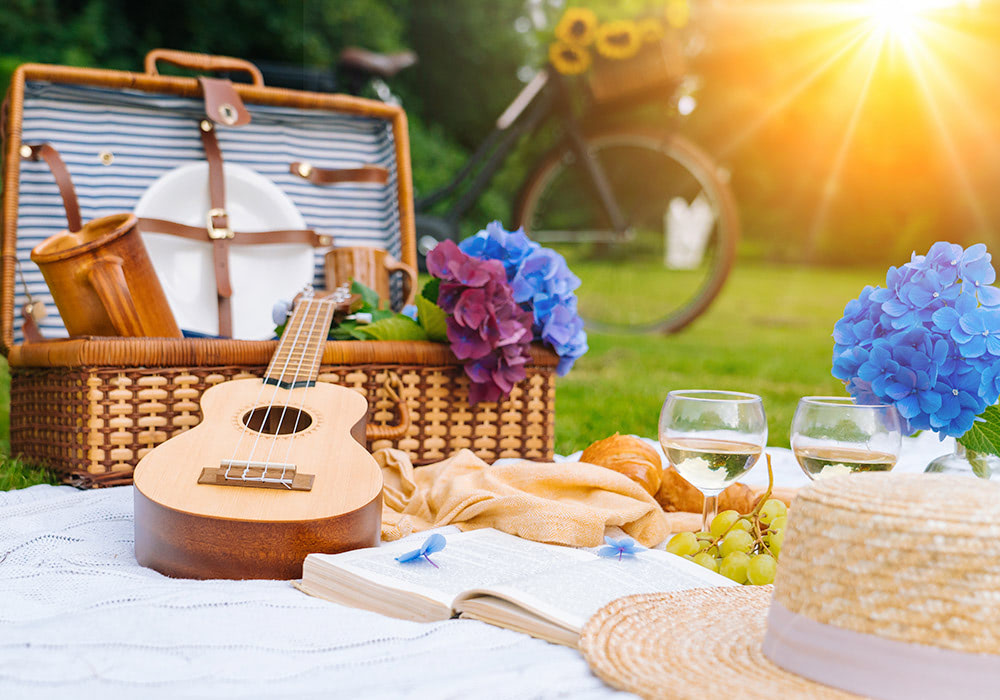 A ukulele rests on a picnic blanket near an open wicker basket, surrounded by flowers, wine glasses, and a sun hat, with a bicycle and sunlight in the background.