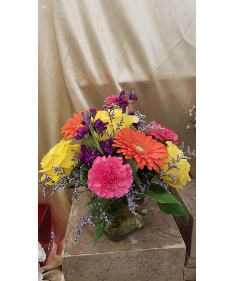 Bright floral arrangement with hot pink carnations, orange gerbera daisies, yellow roses, and purple stock in a glass cube vase.