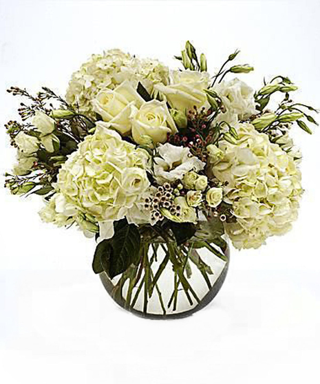 A luxurious floral arrangement of ivory roses, white hydrangeas, and lisianthus, set in a clear vase with lush greenery accents.