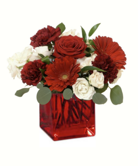 A romantic floral centerpiece featuring red roses, burgundy carnations, red gerbera daisies, ivory spray roses, and flowing greenery like eucalyptus and ruscus.