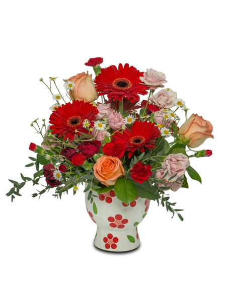 A bright floral arrangement with red Gerbera daisies, peach roses, and pink, white, and red accents, arranged in a whimsical red daisy-patterned pot.