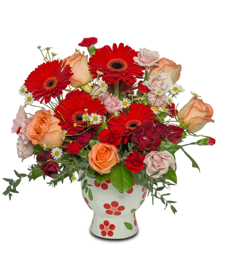 A bright floral arrangement with red Gerbera daisies, peach roses, and pink, white, and red accents, arranged in a whimsical red daisy-patterned pot.