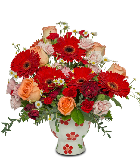 A bright floral arrangement with red Gerbera daisies, peach roses, and pink, white, and red accents, arranged in a whimsical red daisy-patterned pot.