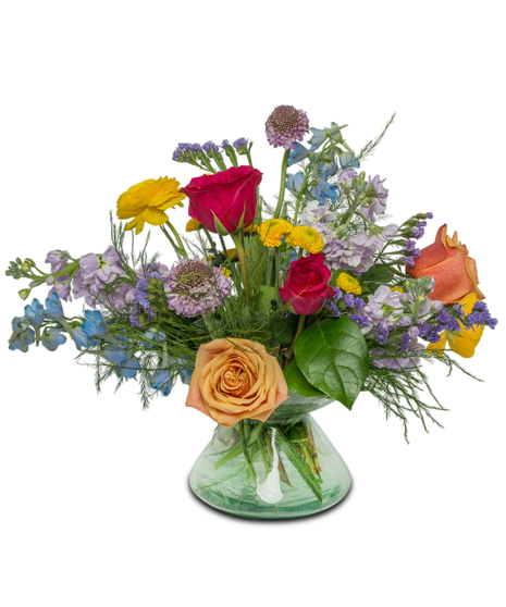 A glass compote vase filled with buttercup ranunculus, roses, fragrant stock, scabiosa, and blue delphinium, arranged in a whimsical, garden-inspired design.