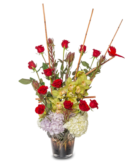 KaBoom floral arrangement with hydrangeas, red roses, cymbidium orchids, bamboo sticks, and a red cardinal in a hand-blown glass vase with rose accents and a tinted base.