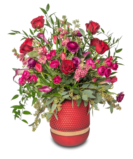 Lovespell floral arrangement featuring red roses, ranunculus, calla lilies, fragrant stock, tulips, and spray roses in a textured red vase with gold accents.