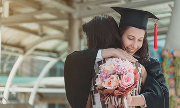 A graduate in cap and gown hugs someone while holding a bouquet of flowers in an outdoor setting with architectural structures in the background.