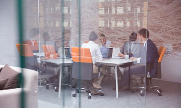 People are sitting around a conference table, engaged in a meeting, inside a modern glass-walled office with city buildings visible through the windows.