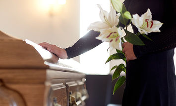 Person in black clothing holding white lilies, standing next to a wooden coffin in a dimly lit room, creating a somber atmosphere.