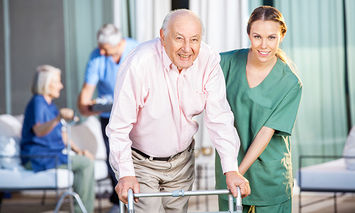 An elderly man using a walker is assisted by a smiling female healthcare worker in green scrubs. Other seniors are visible in the background, also receiving care in an outdoor setting.